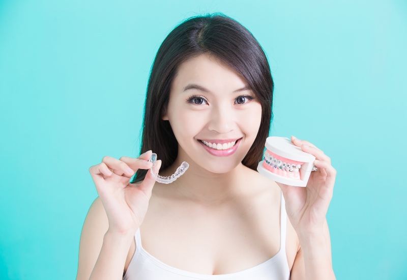 A woman compares Invisalign and traditional braces, holding an aligner & braces to show pros & cons